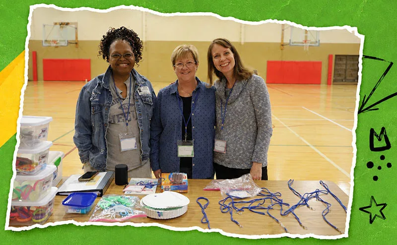 Three club volunteers stand behind a table of activity supplies in a school gym.