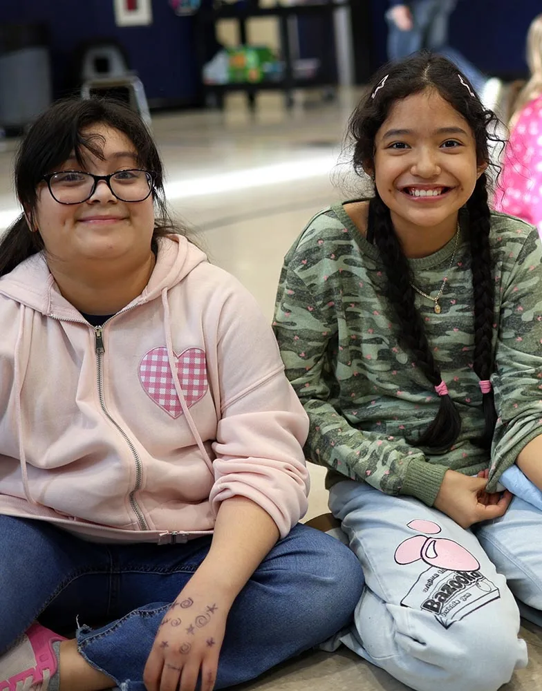 what-happens-at-kids-club Two girls smile together while seated in a school gym during Crossroads Kids Club.